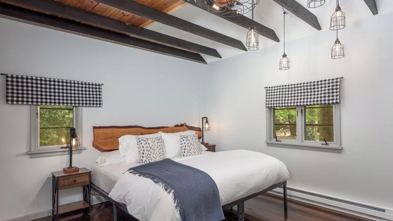 Une chambre à coucher comprenant un lit, deux tables de chevet, une tête de lit et deux fenêtres ornées de rideaux à carreaux.