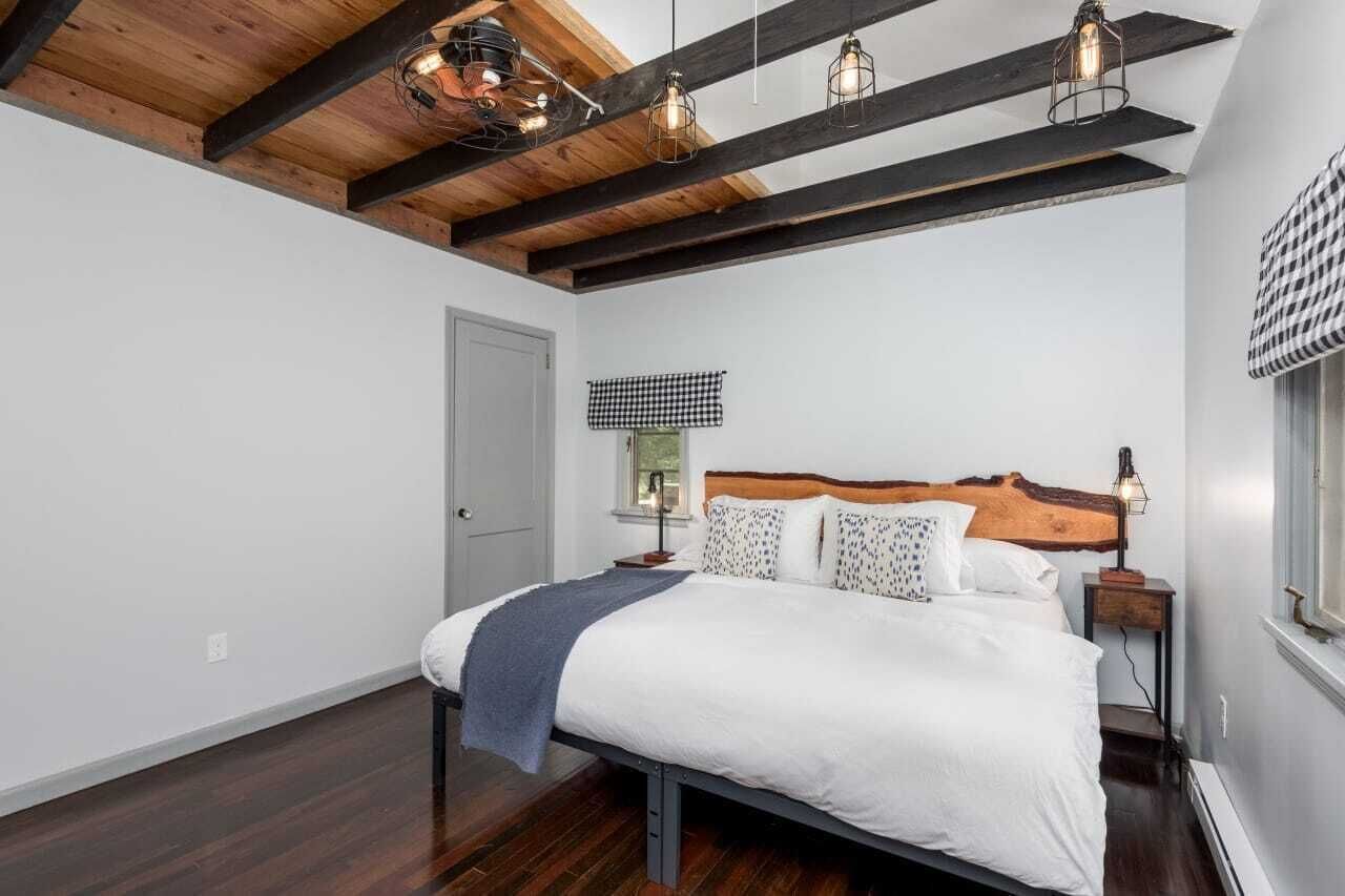 Un dormitorio con techo de madera, una cama con ropa blanca, un visillo de ventana a cuadros y una mesita de noche con una lámpara.