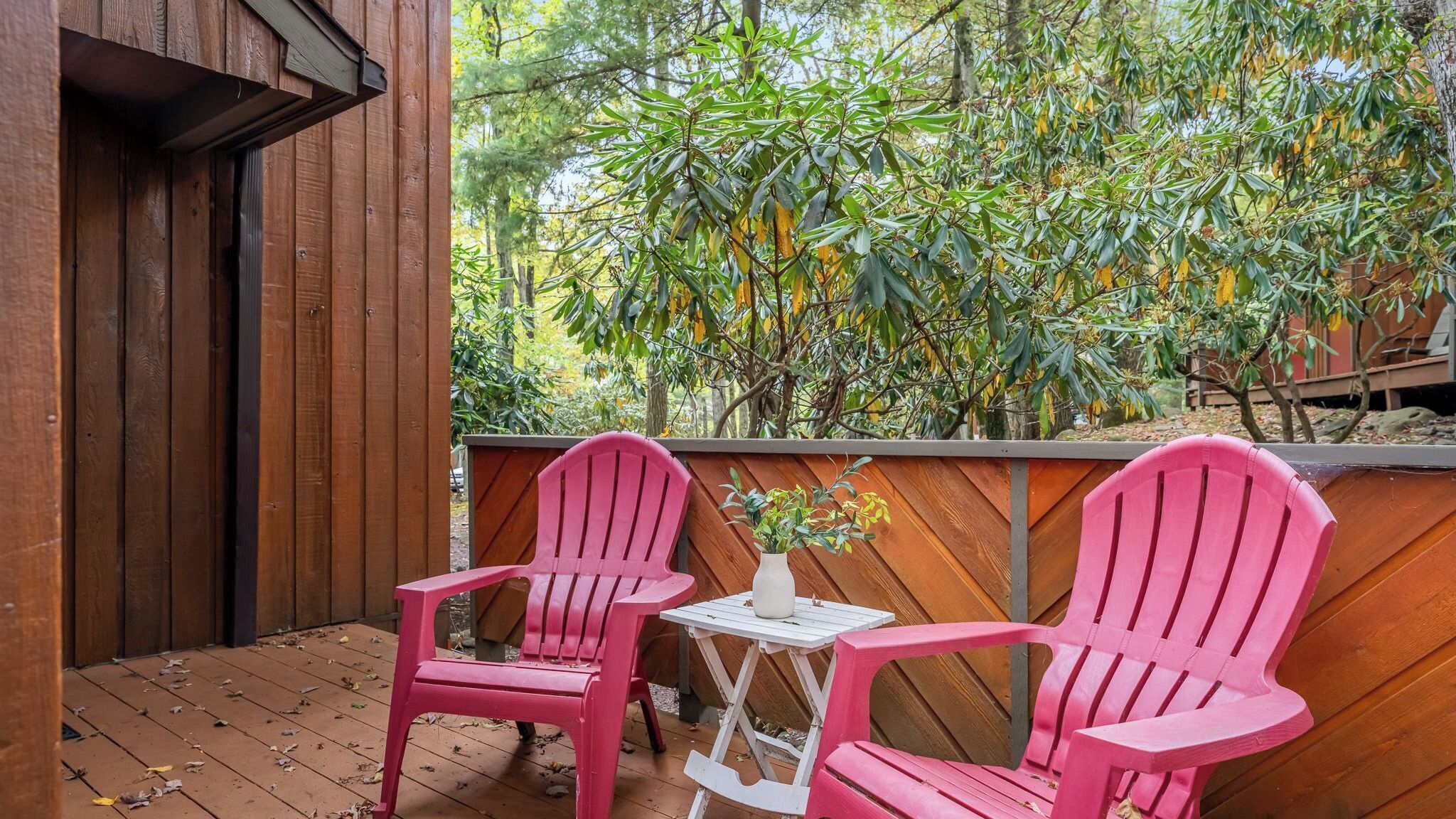Una terraza de madera con dos sillas rosas y una mesita, rodeada de abundante vegetación.