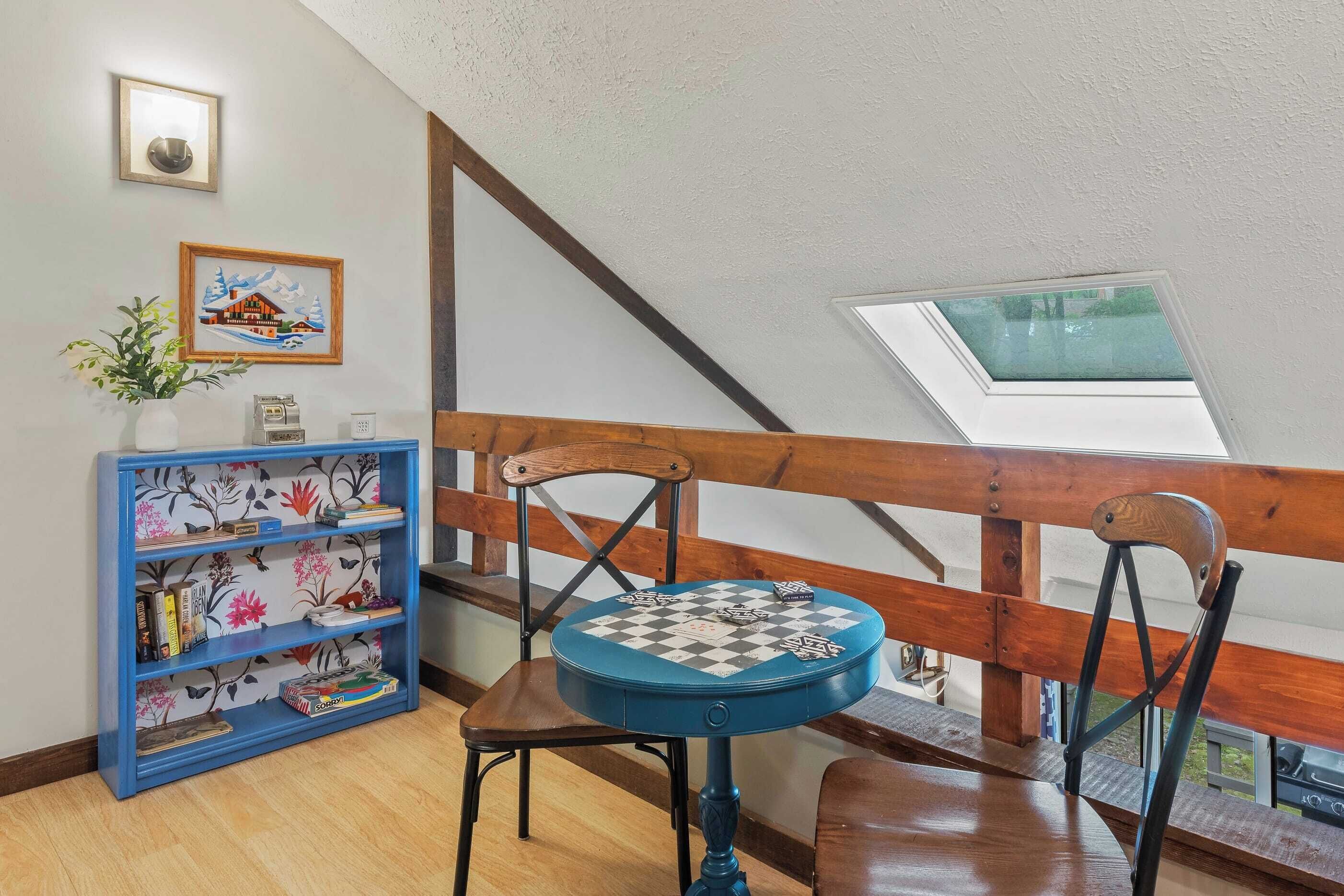 Een kamer met een houten trap, een blauwe boekenkast, een ronde tafel met een schaakbord en een dakraam.