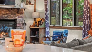 A cozy living room with a fireplace, a comfortable chair with a 'WELCOME TO THE LAUNCH' pillow, and a view of greenery outside.