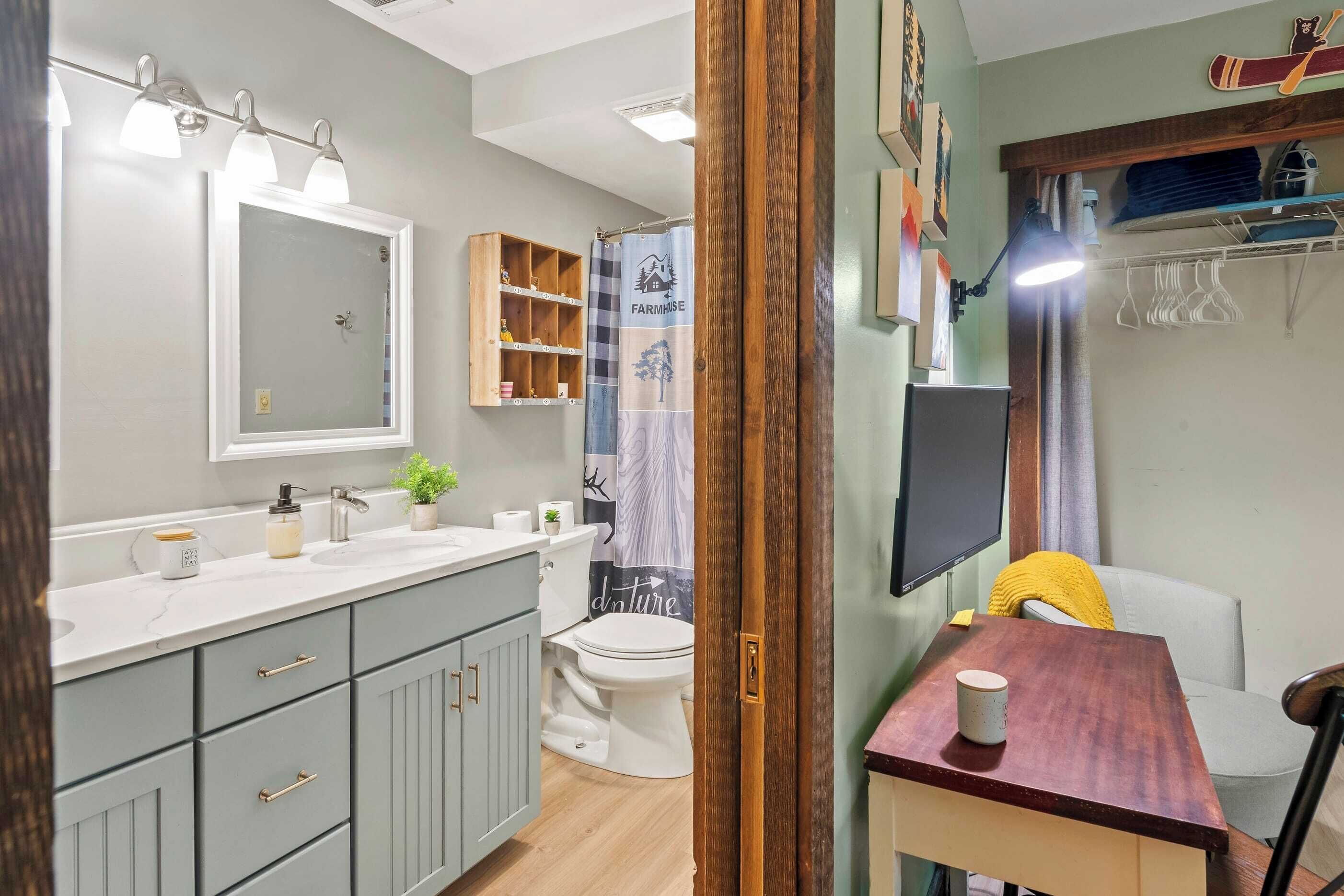 Une salle de bain avec un lavabo blanc, un miroir et des toilettes, un rideau de douche au motif champêtre et une étagère en bois où sont rangés les articles de toilette.