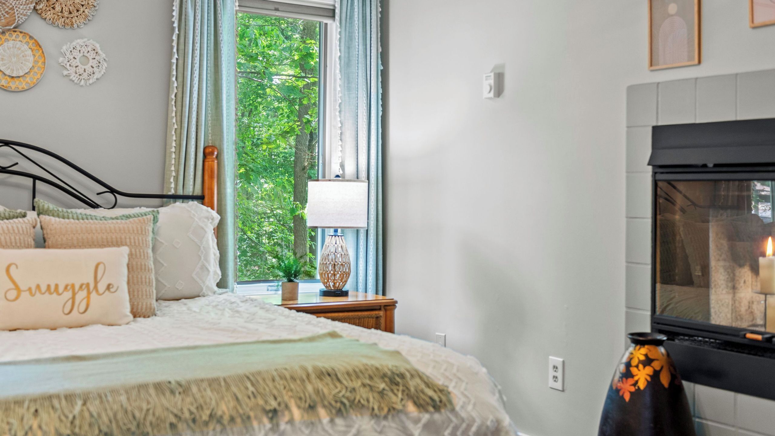 A bedroom with a bed, pillows, a nightstand, a lamp, a window with curtains, and a fireplace.