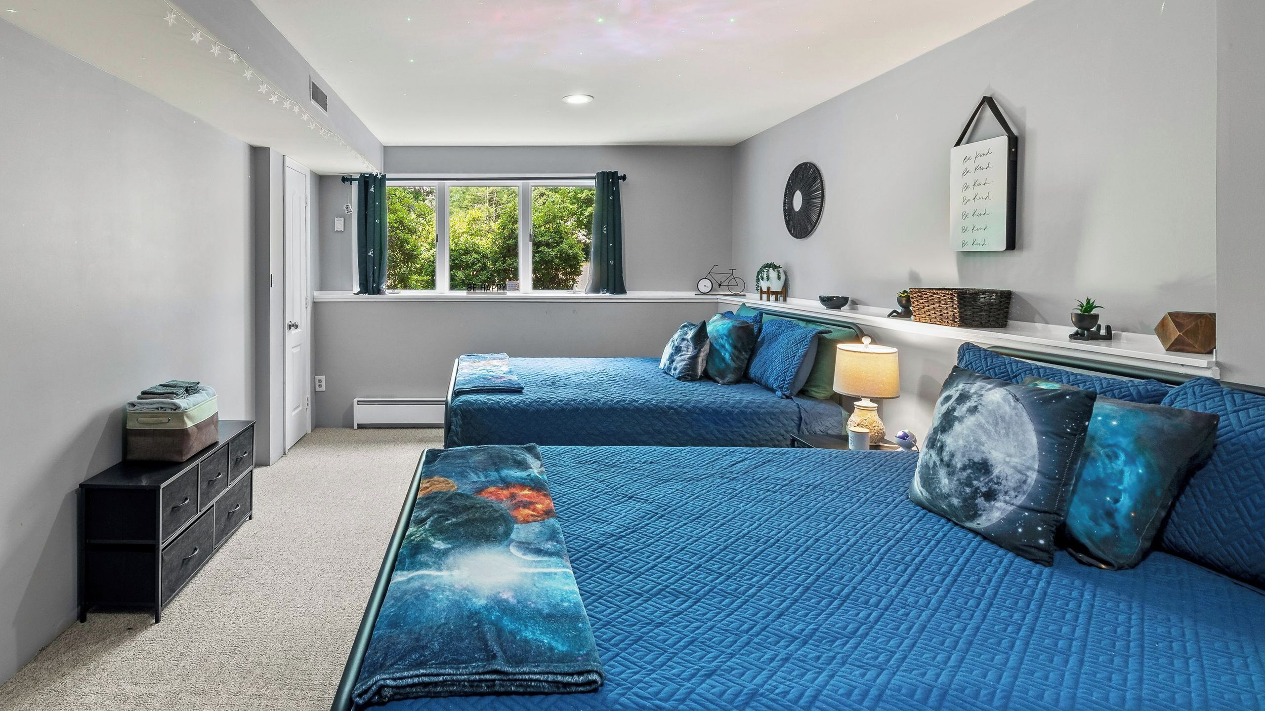 Un dormitorio con dos camas, una ventana con cortinas, un reloj de pared, un cartel motivacional y una pequeña estantería con adornos.