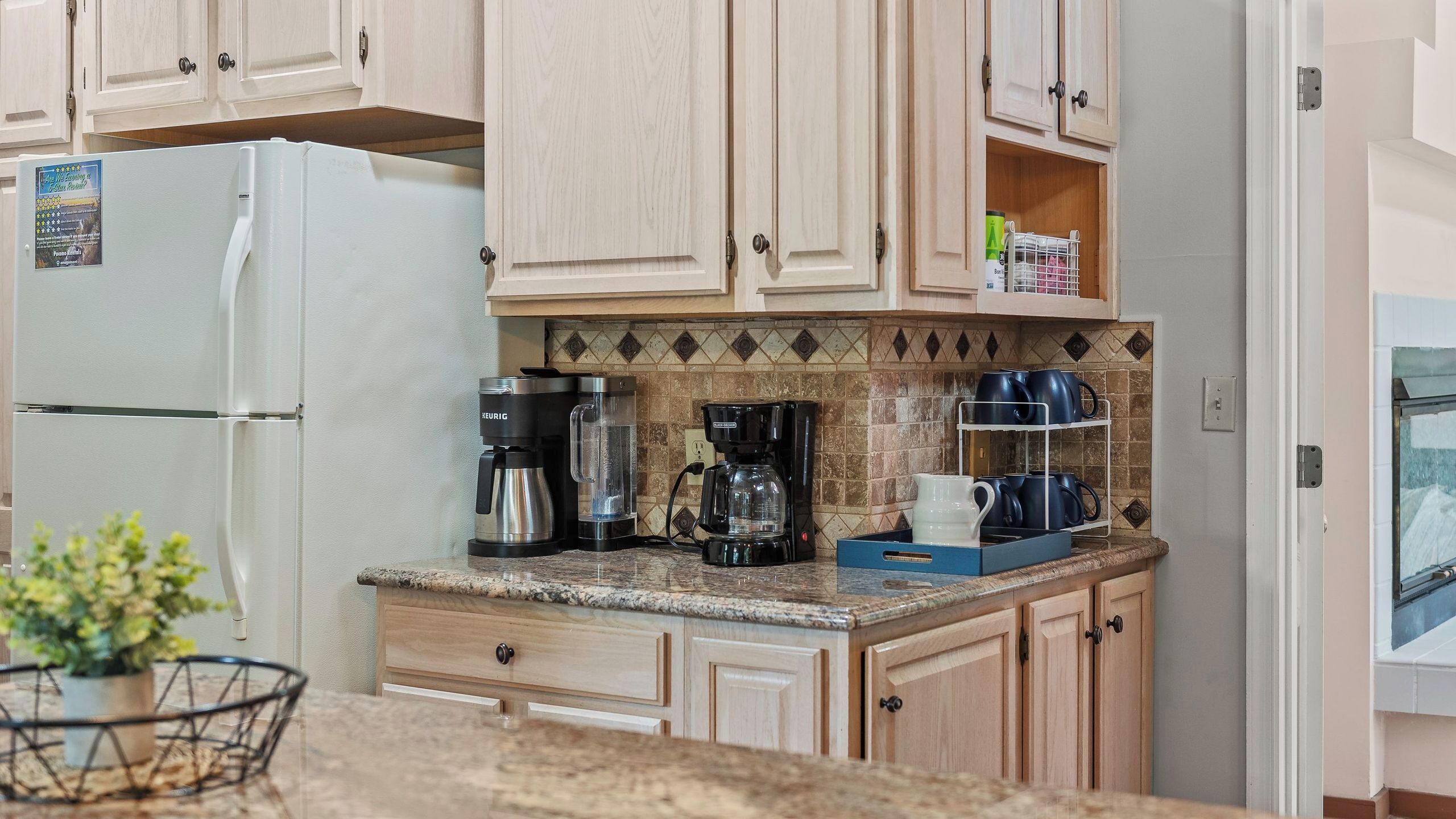 A kitchen with a refrigerator, coffee maker, and a shelf with cups.