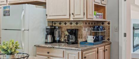 A kitchen with a refrigerator, coffee maker, and a shelf with cups.