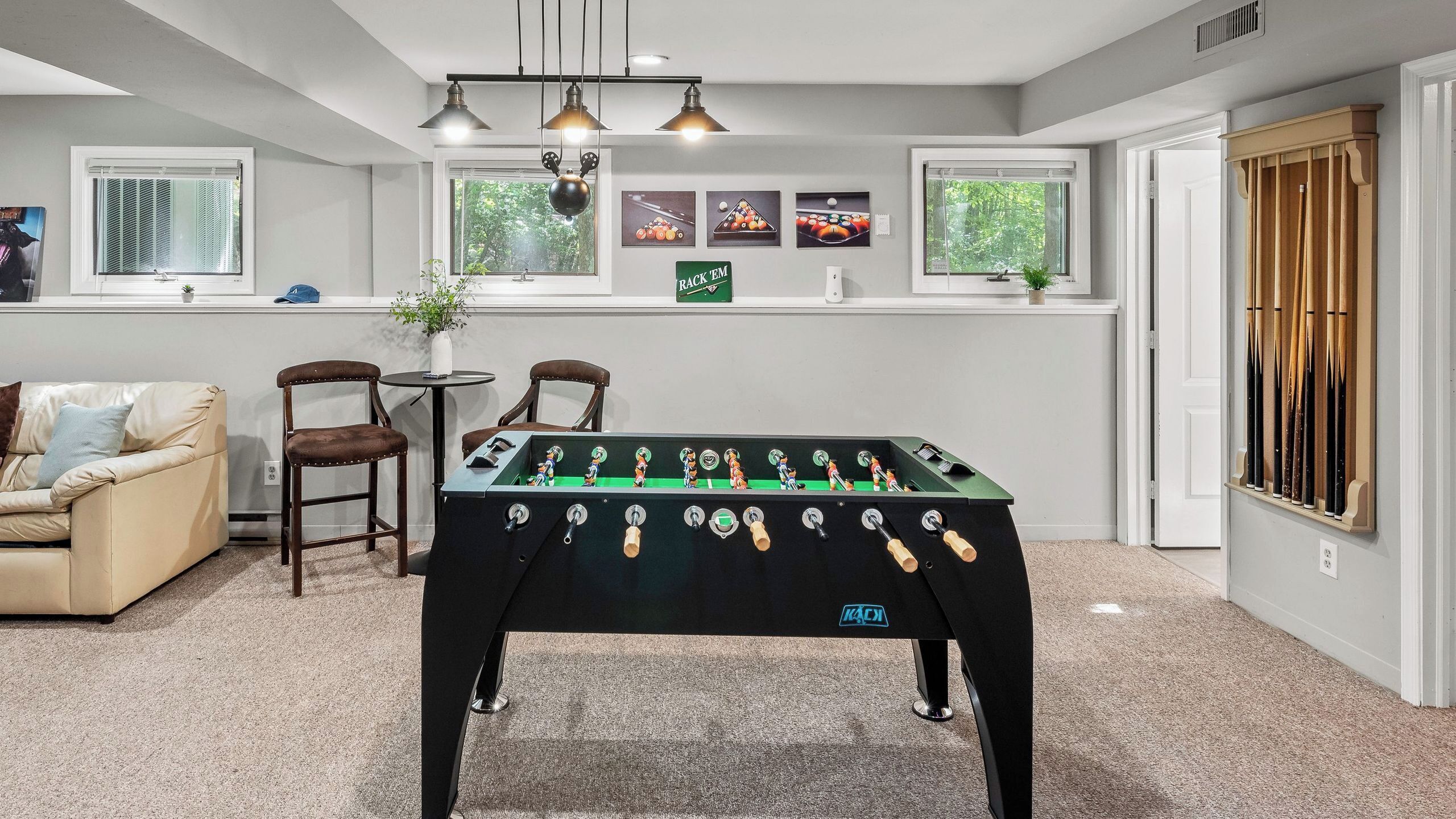 A room with a foosball table, a sofa, two chairs, a small table with a plant, and a window with blinds.