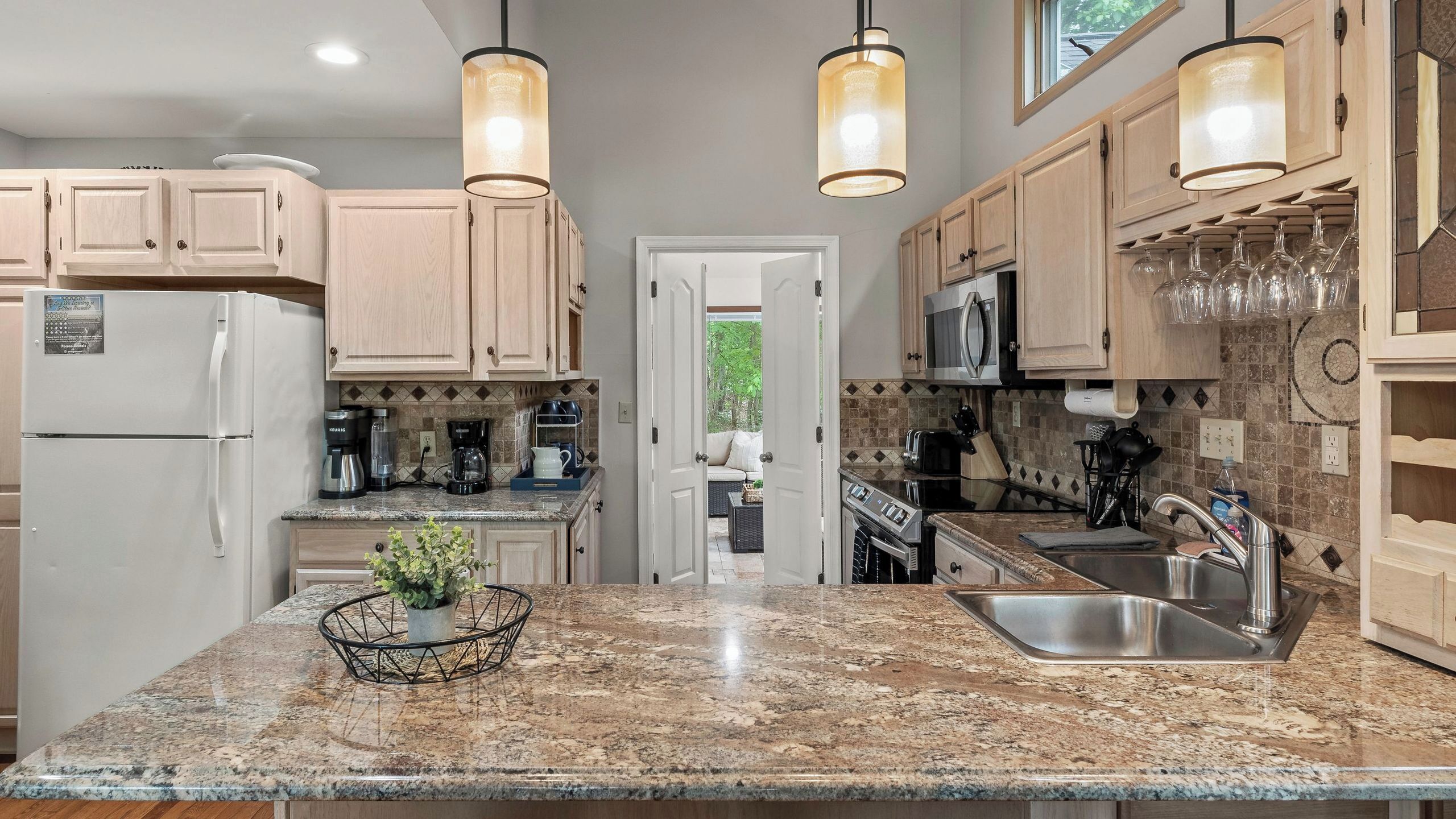 A kitchen with granite countertops, wooden cabinets, a refrigerator, a microwave, a sink, and pendant lighting.