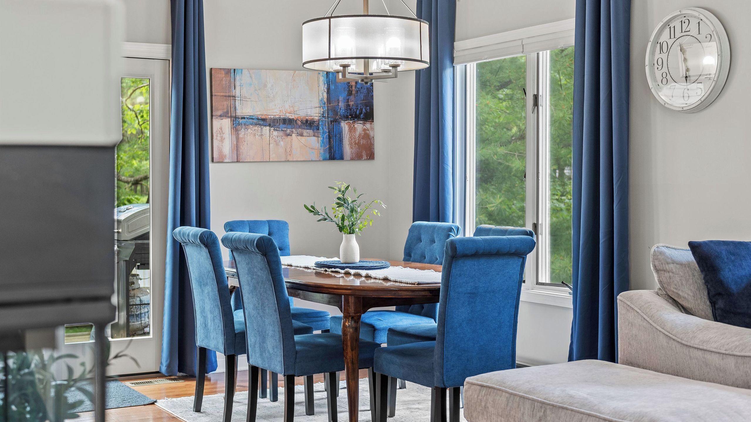 A dining area with a round table, blue upholstered chairs, a vase with greenery, and a large abstract painting on the wall.