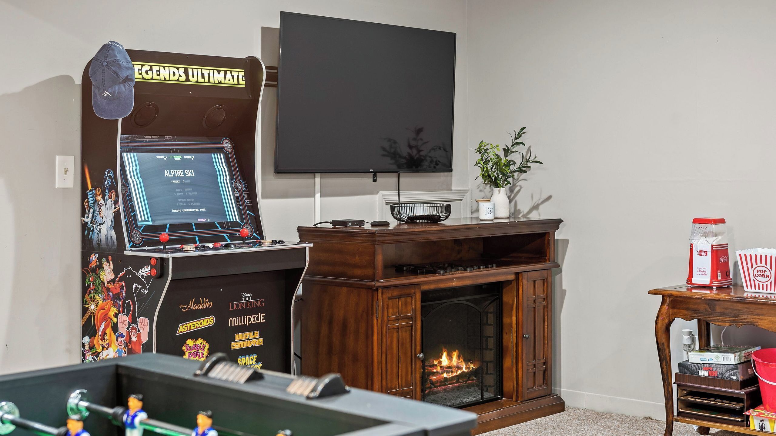 A room with a foosball table, a fireplace, a TV, and a wall-mounted screen displaying a game.