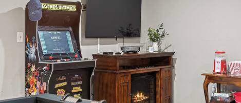 A room with a foosball table, a fireplace, a TV, and a wall-mounted screen displaying a game.