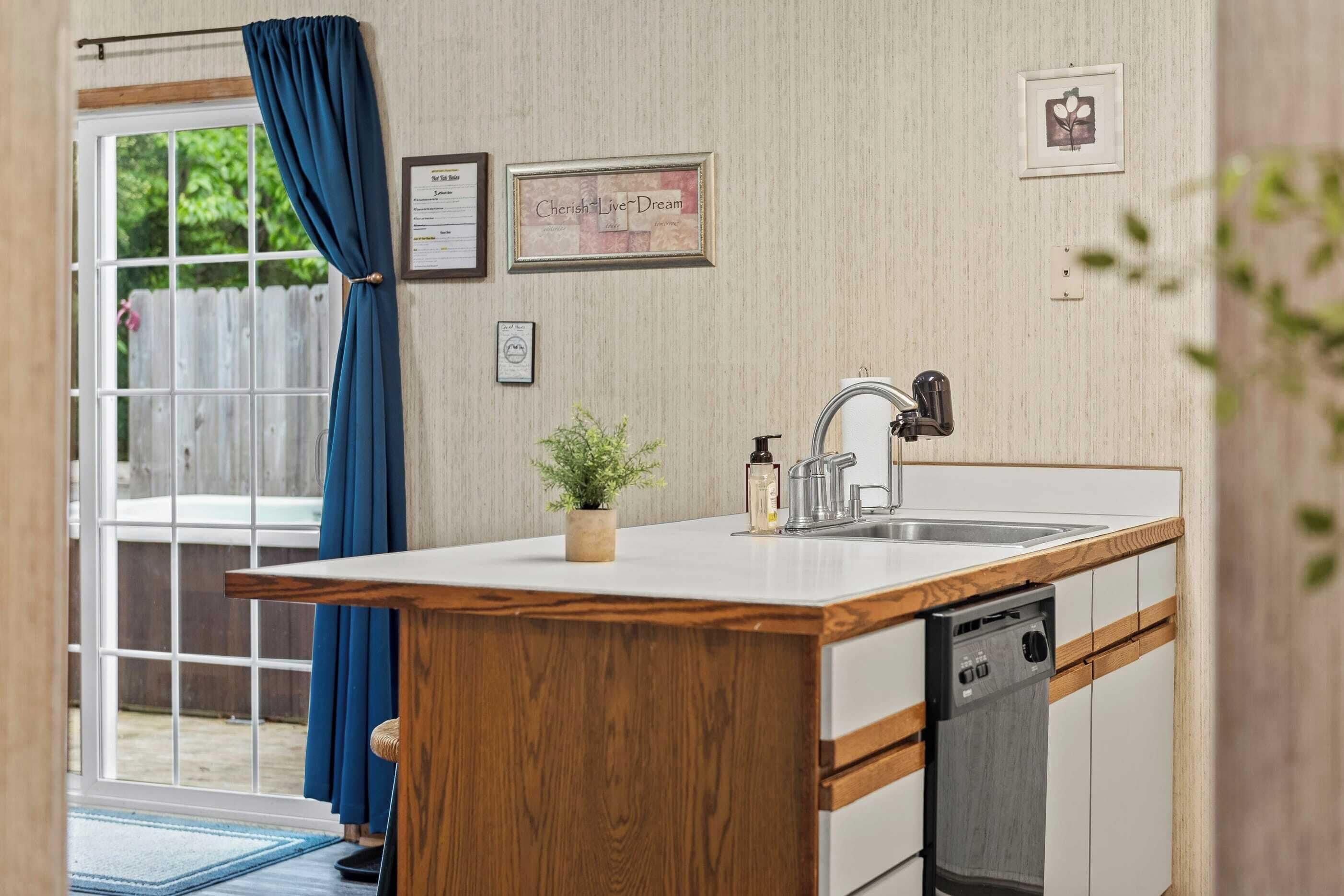 A kitchen with a wooden countertop, a sink, and a dishwasher. There is a window with blue curtains, a small plant on the countertop, and a framed picture on the wall.