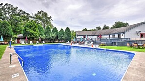 A rectangular swimming pool with a metal ladder, surrounded by lounge chairs and umbrellas, with a building and trees in the background. - Harmony Ridge by Avantstay Fireplace, Large Deck & Forest Views (Lake Harmony)
