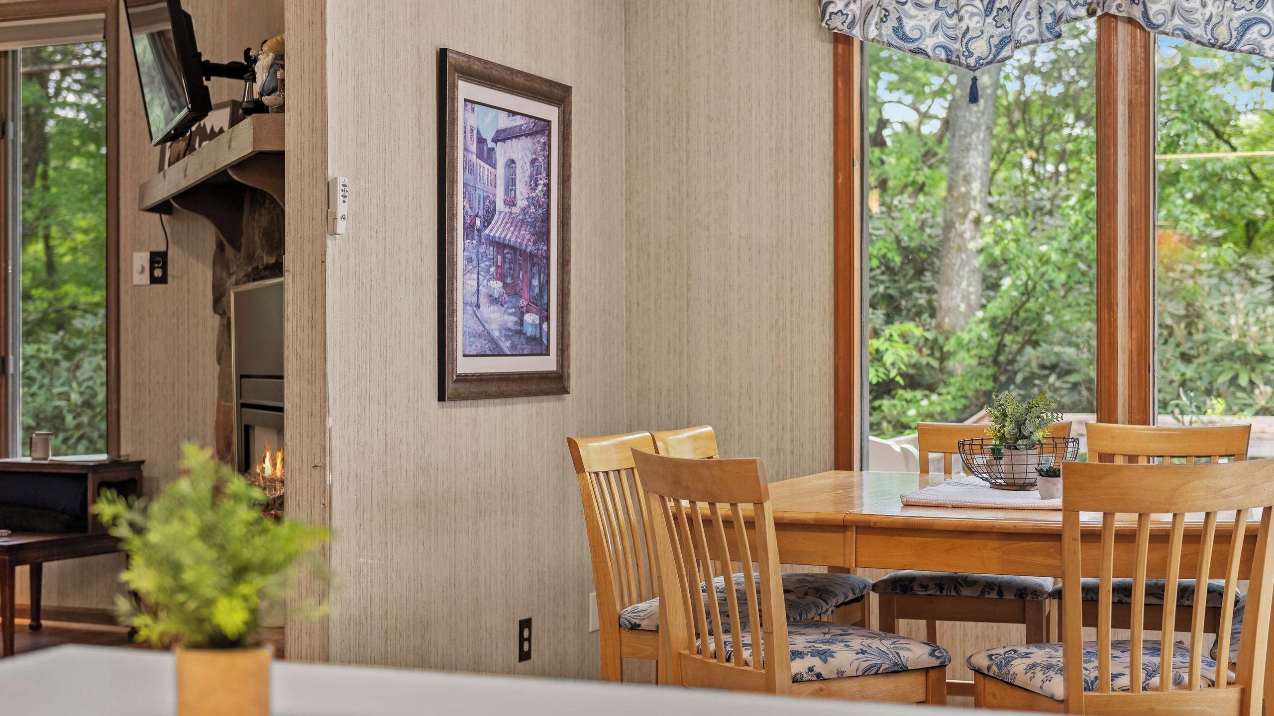 Un coin repas avec des chaises et une table en bois, un tableau encadré au mur et une vue sur la verdure à l’extérieur.