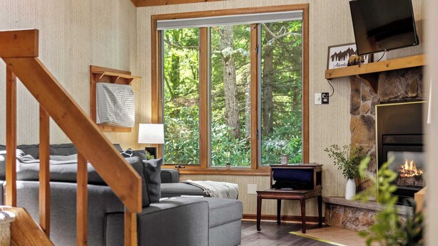 A cozy living room with a large window, a fireplace, a sofa, and a TV. - Harmony Ridge by Avantstay Fireplace, Large Deck & Forest Views (Lake Harmony)