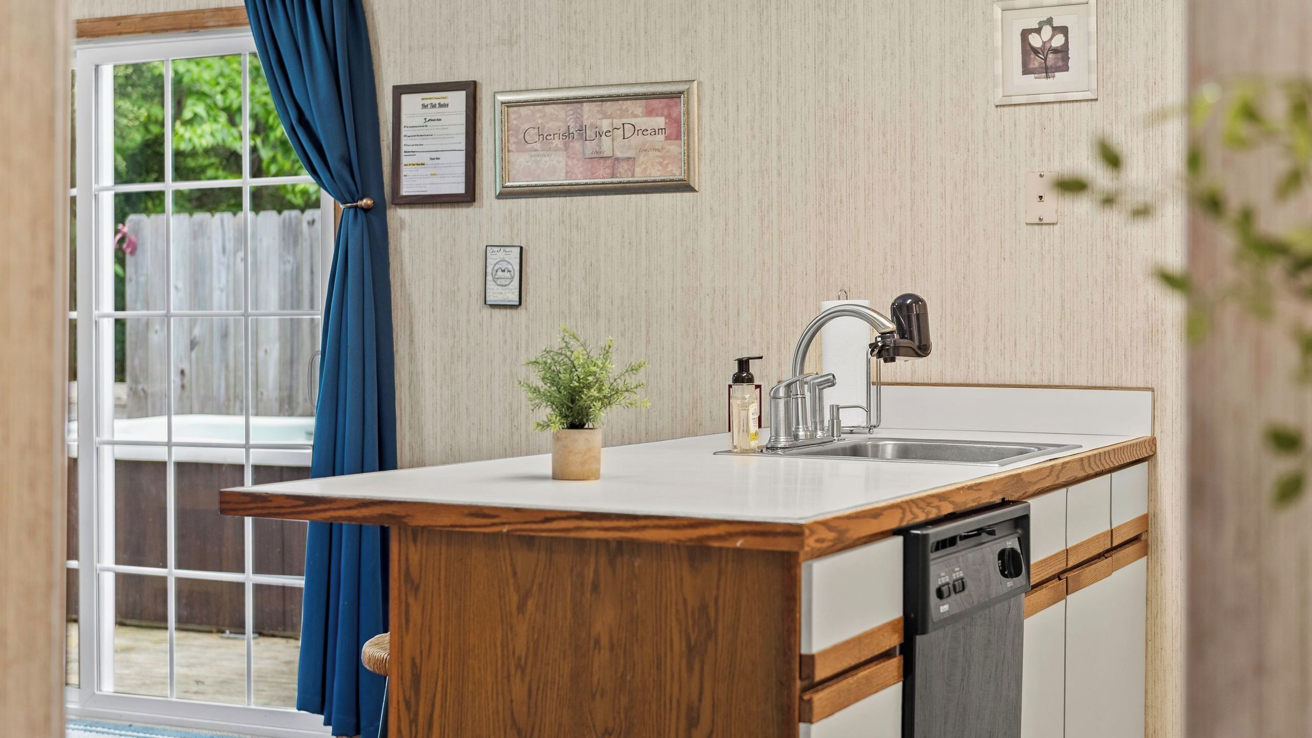 A kitchen with a wooden countertop, a sink, a dishwasher, a potted plant, and a window with blue curtains.