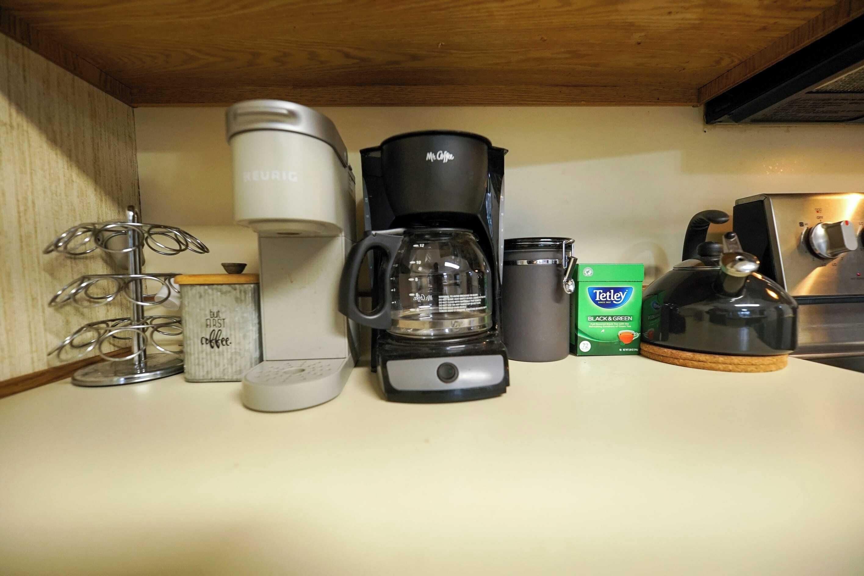 Uma bancada de cozinha com diversos equipamentos para fazer café, incluindo uma cafeteira Keurig, uma cafeteira Mr. Coffee, uma jarra de vidro, uma cafeteira Teflex e uma caixa de saquinhos de chá Tetley.