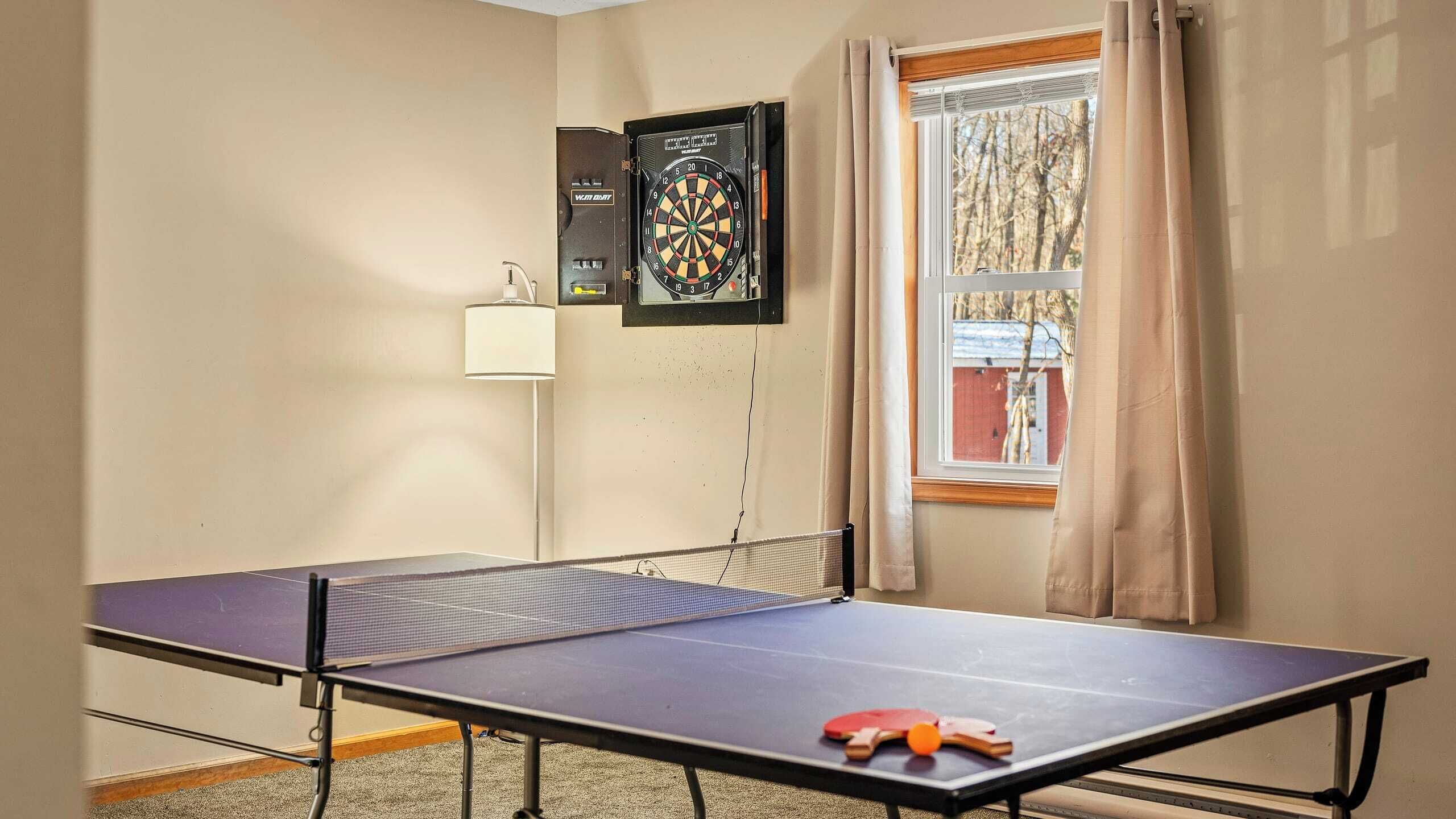 A room with a ping pong table, a dartboard, and a window with curtains.