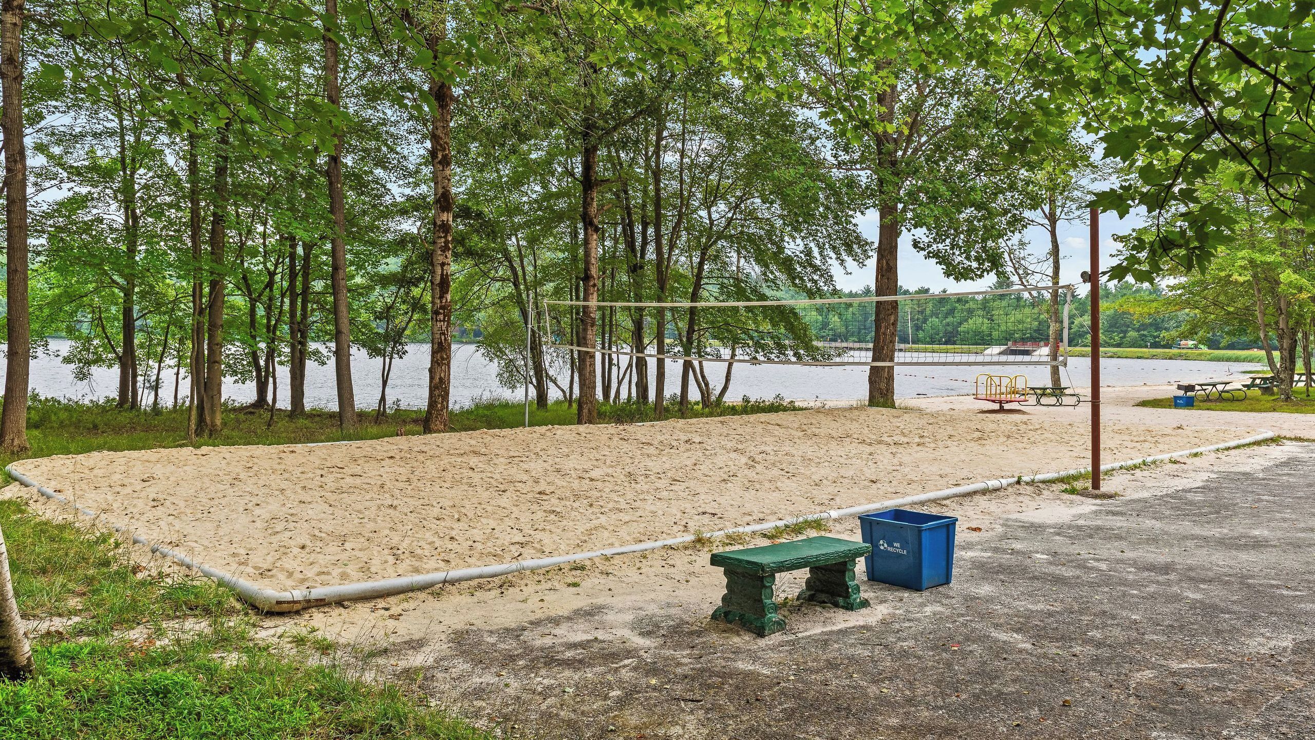 A sandy beach area with a volleyball net, a green bench, and a blue recycling bin.