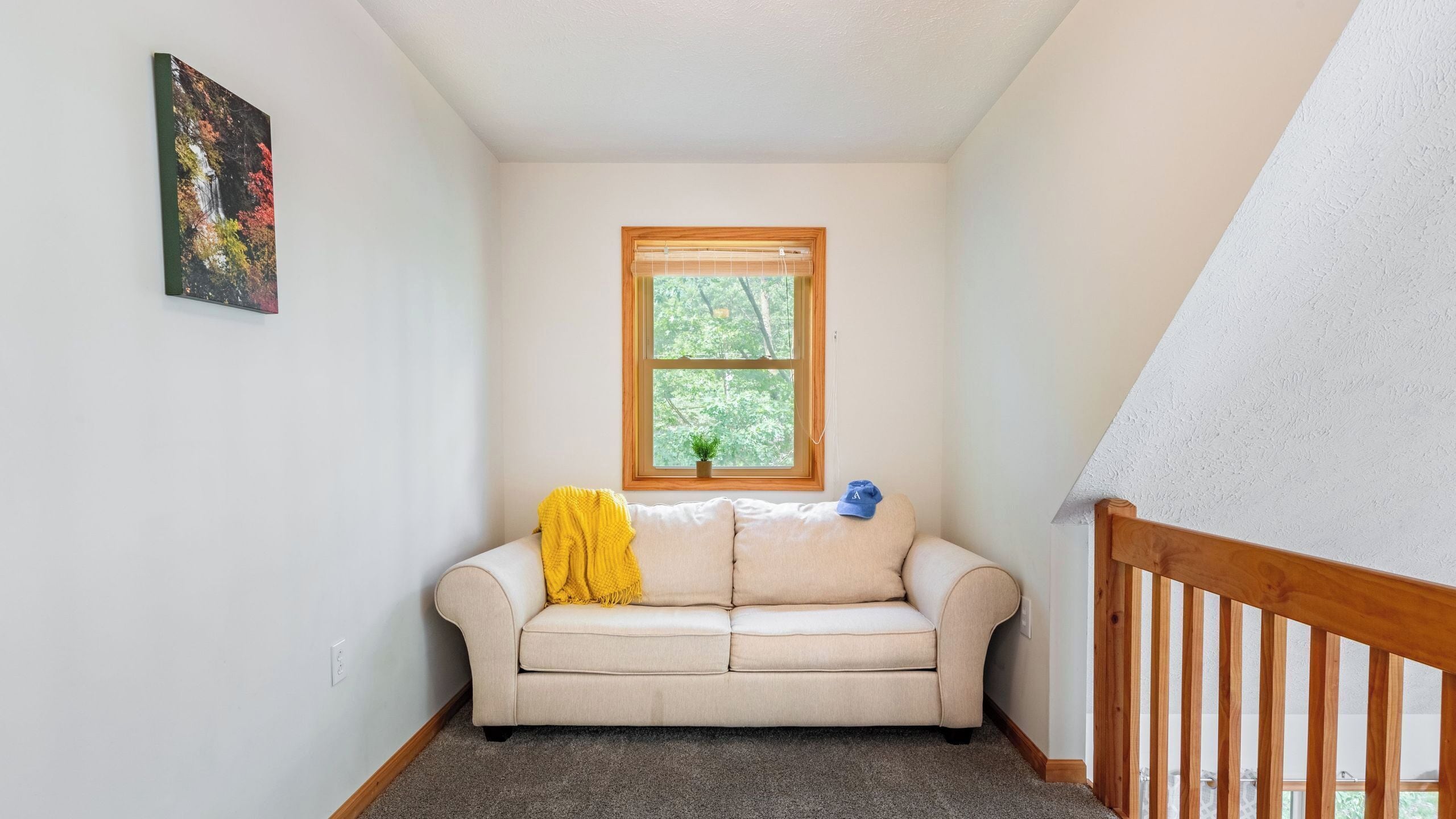 A room with a beige sofa, a small window with a view of trees, a painting on the wall, and a wooden railing.