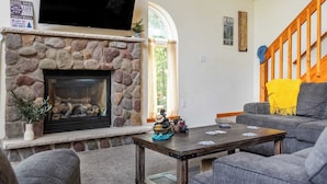 A cozy living room with a stone fireplace, a flat-screen TV mounted above it, and a comfortable seating area with a gray sofa and a wooden coffee table. - The Nest at Brier Crest by Avantstay Game Room, Expansive Deck & Fire Pit (Blakeslee)