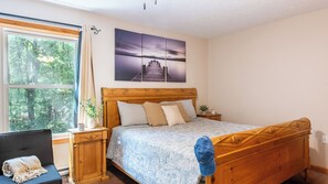 Un dormitorio con cama de madera, un escritorio, una silla, una ventana con vista a árboles y un cuadro en la pared.