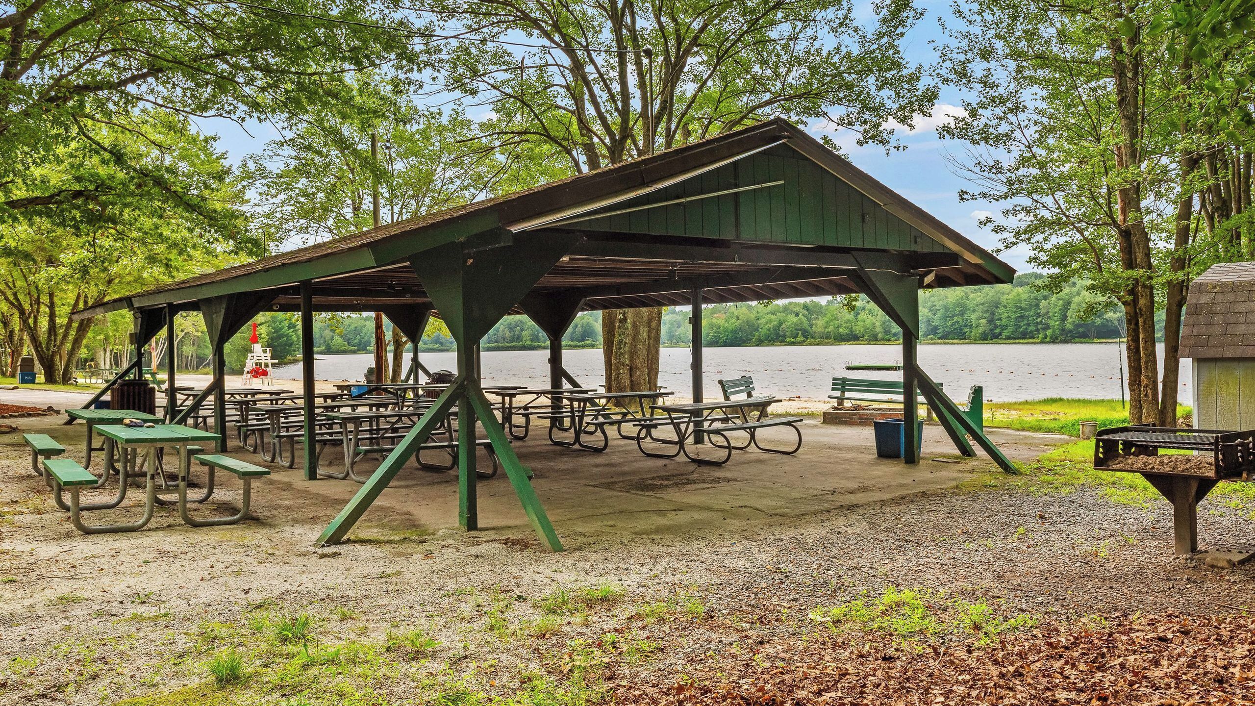 A covered picnic area with benches and picnic tables, surrounded by trees and a lake.