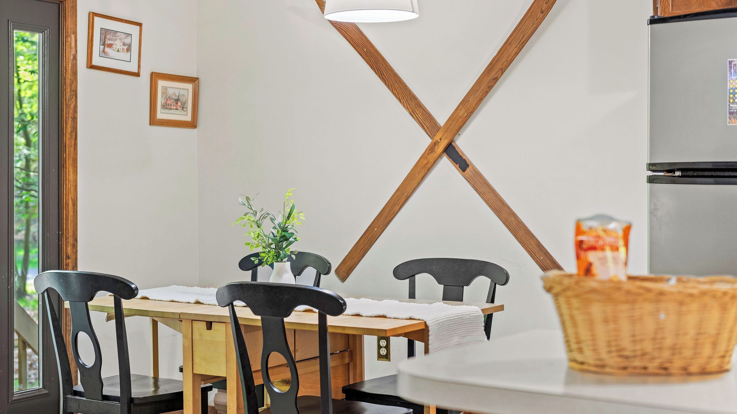 Un coin repas avec une table en bois, des chaises noires, une nappe blanche, une plante en pot et un réfrigérateur.