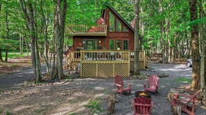 A cabin with a deck, surrounded by trees, and a fire pit with chairs.