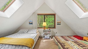 A bedroom with a sloped ceiling, a bed with a plaid bedspread, a bedside table with a lamp, a small shelf with books, and a window with a view of trees. - 7mins to Skiing, Fire Pit, Game Table Lake Access Hygge Home by Avantstay (Blakeslee)