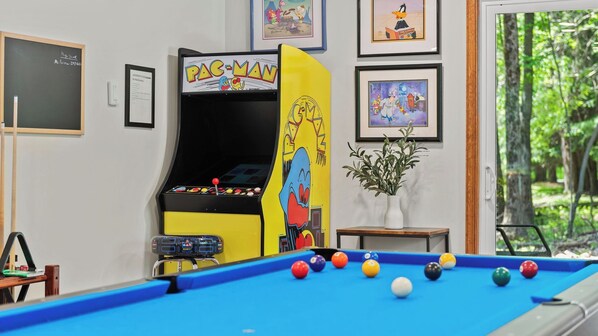 Una mesa de billar con bolas de varios colores, una máquina de Pac-Man, obras de arte enmarcadas en la pared y una ventana con vistas a árboles.