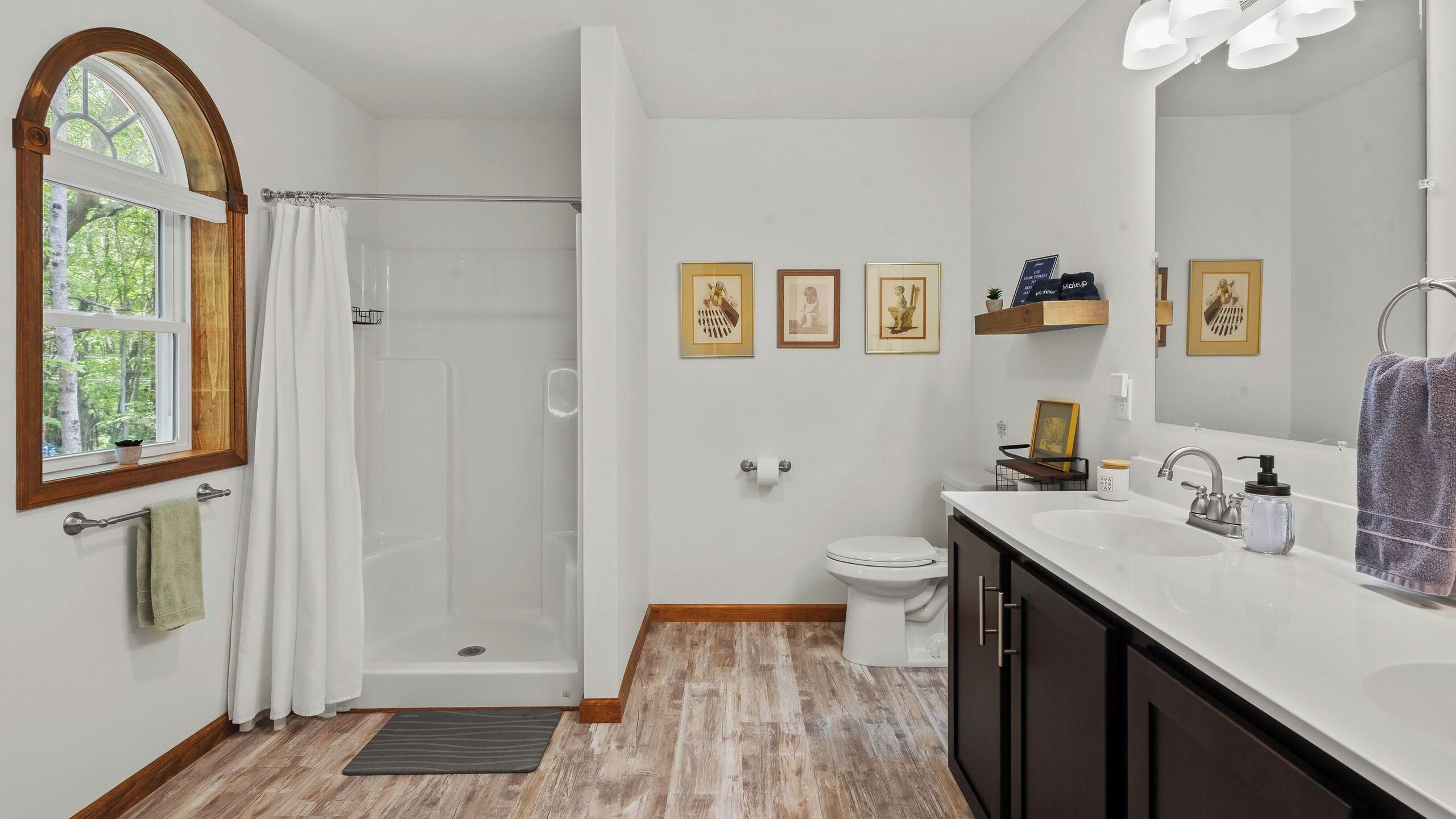 Et moderne badeværelse med bruser, toilet, vask og underskab. Der er billeder i rammer på væggen, et vindue med udsigt til træer og en bøjle til håndklæder.