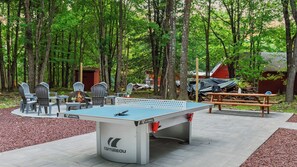 Ein Tischtennistisch im Freien mit Stühlen, ein Picknicktisch und eine Feuerstelle in bewaldeter Umgebung.