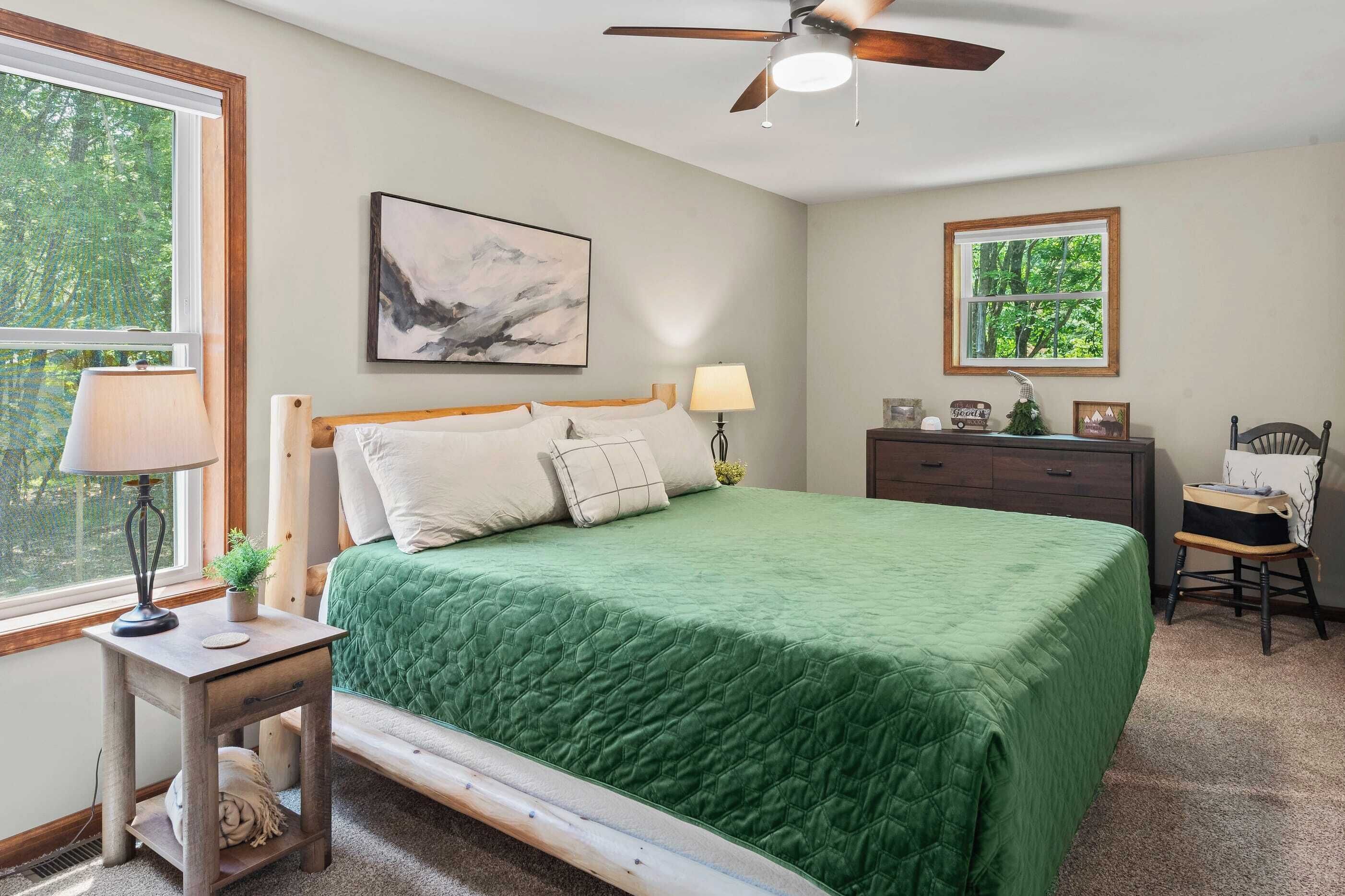 A bedroom with a bed, a nightstand, a desk, a chair, a ceiling fan, a window, and a framed picture on the wall.