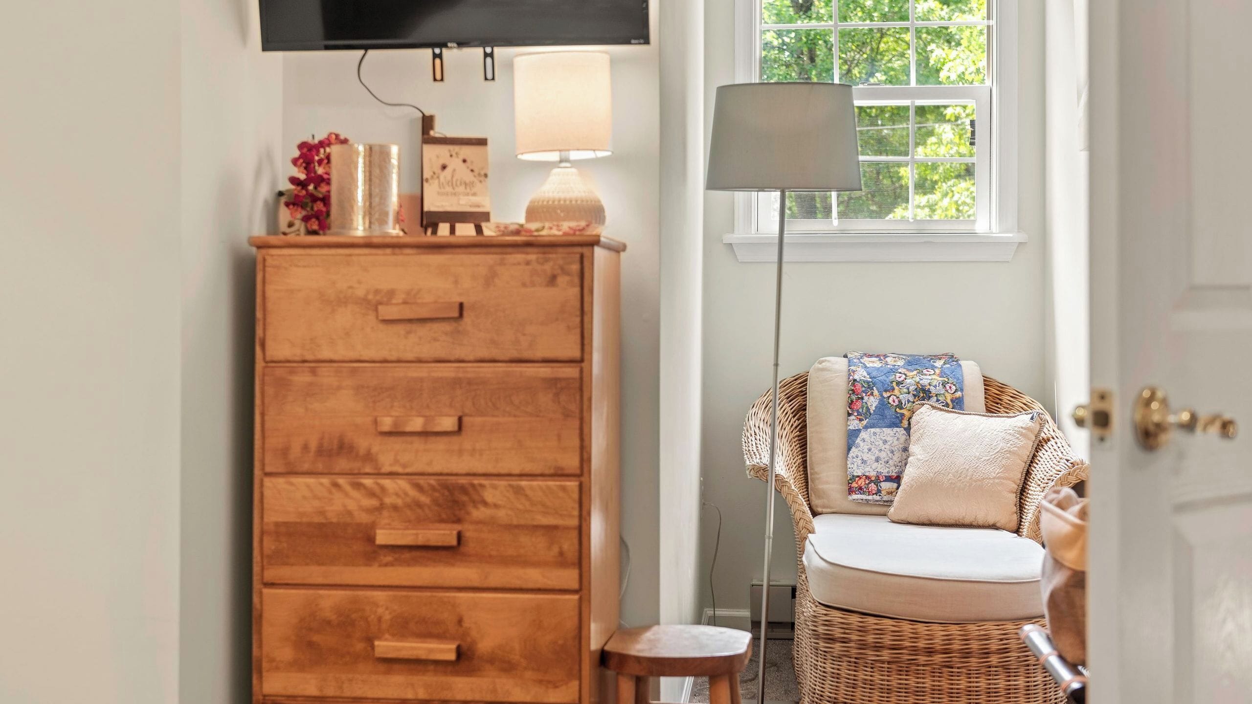 Une commode en bois avec des tiroirs, une chaise en osier avec des coussins, un lampadaire et un téléviseur fixé au mur.