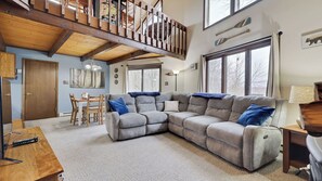 A spacious living area with a large sectional sofa, a dining table with chairs, a staircase, and large windows. - Chalet All Day Avantstay Lakeside, Firepit, Shared Pool (Pocono Lake)