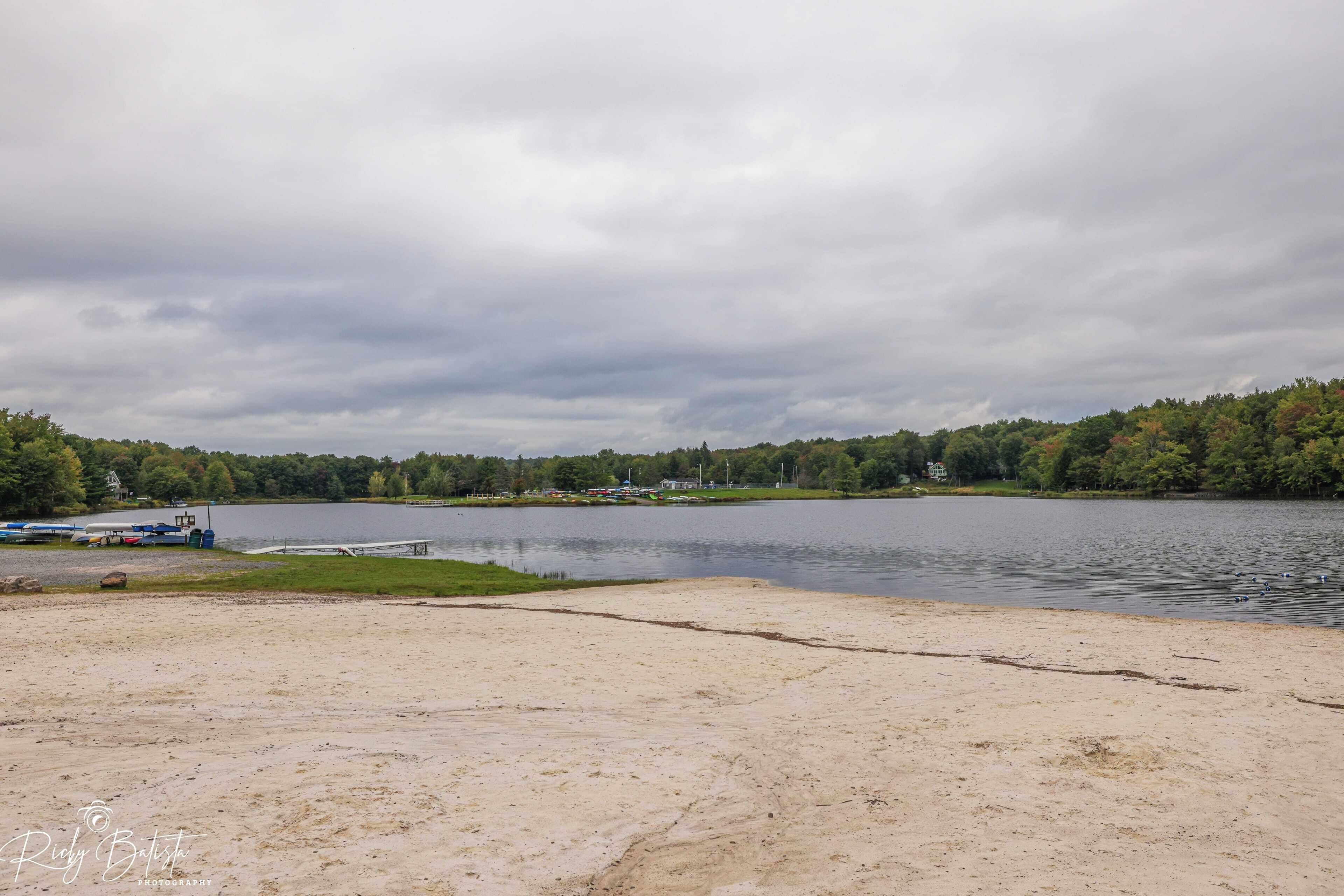 Udsigt over en sø med sandstrand, en anløbsbro og et skovområde i baggrunden.