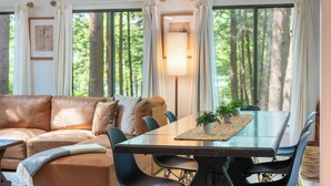 Ein Wohnzimmer mit einem braunen Ledersofa, einem Glastisch mit Stühlen, einer Stehlampe und Blick auf Bäume durch große Fenster.