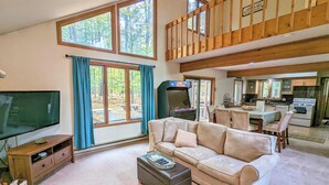 A spacious living room with a large television, a sofa, and a coffee table. There is a kitchen area with a dining table and chairs visible through an open door.