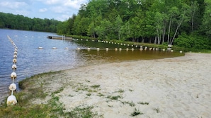 A sandy beach with a row of buoys and a dock extending into a lake surrounded by dense forest.