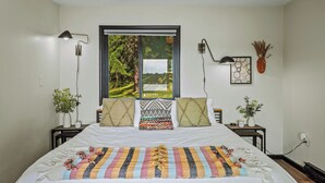 A bedroom with a large bed, two bedside tables, a window with a view of greenery, and wall-mounted lamps.