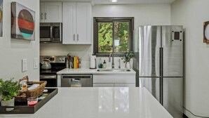 A modern kitchen with white cabinets, stainless steel appliances, a window with a view of trees, and a center island with a basket and a glass.