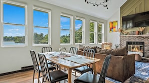 A room with a dining table, chairs, a sofa, a fireplace, and large windows with a view of trees and a lake. - Strawberry Shores by Avantstay Lakefront Private Beach (Long Pond)