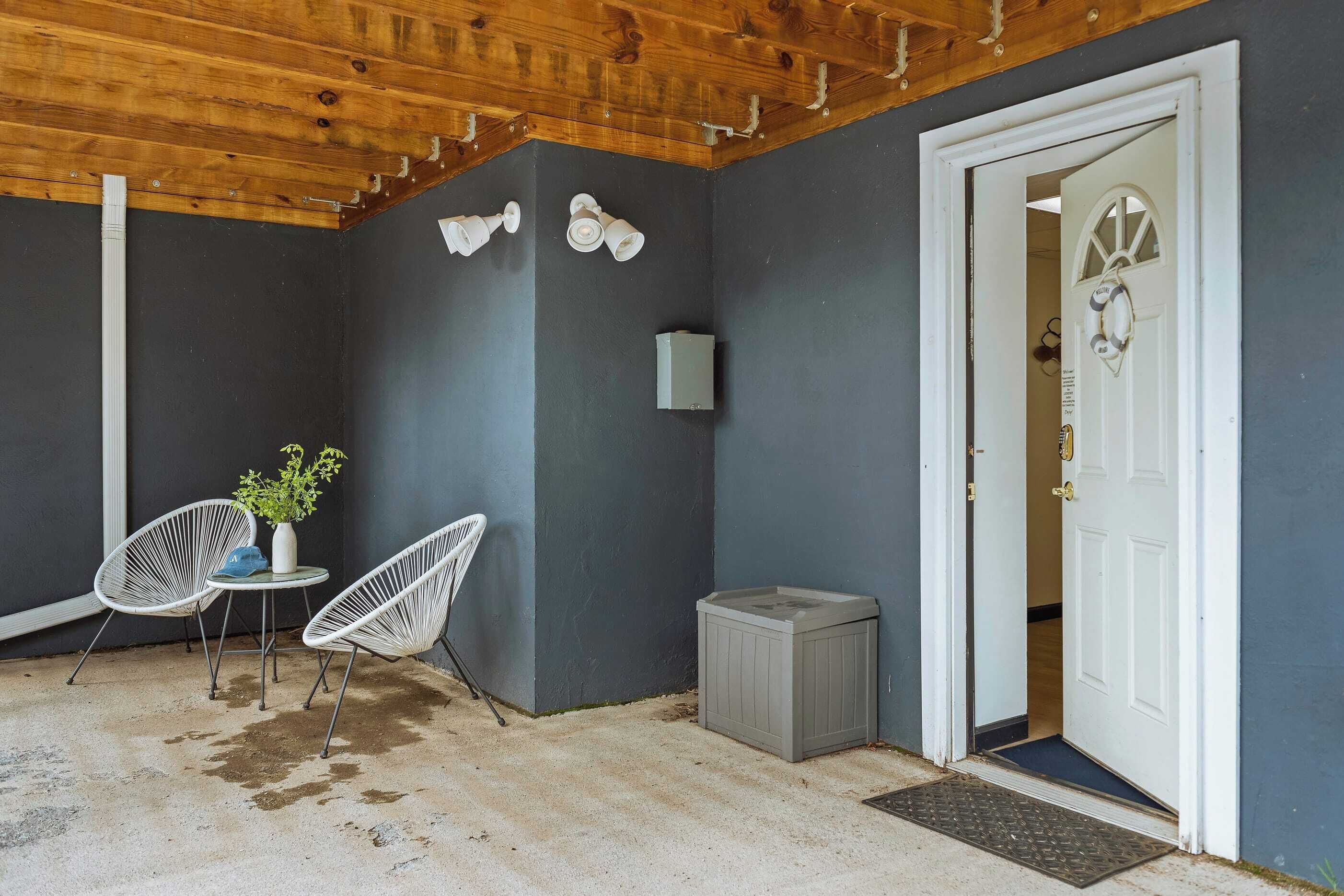 Una habitación con pared gris oscura, techo de madera, puerta blanca con picaporte decorativo, una mesita blanca con planta en maceta y dos sillas blancas.