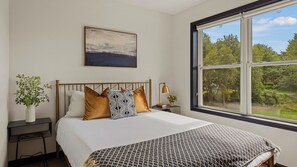 A bedroom with a bed, a bedside table, a lamp, a window with a view of trees, and a painting on the wall.