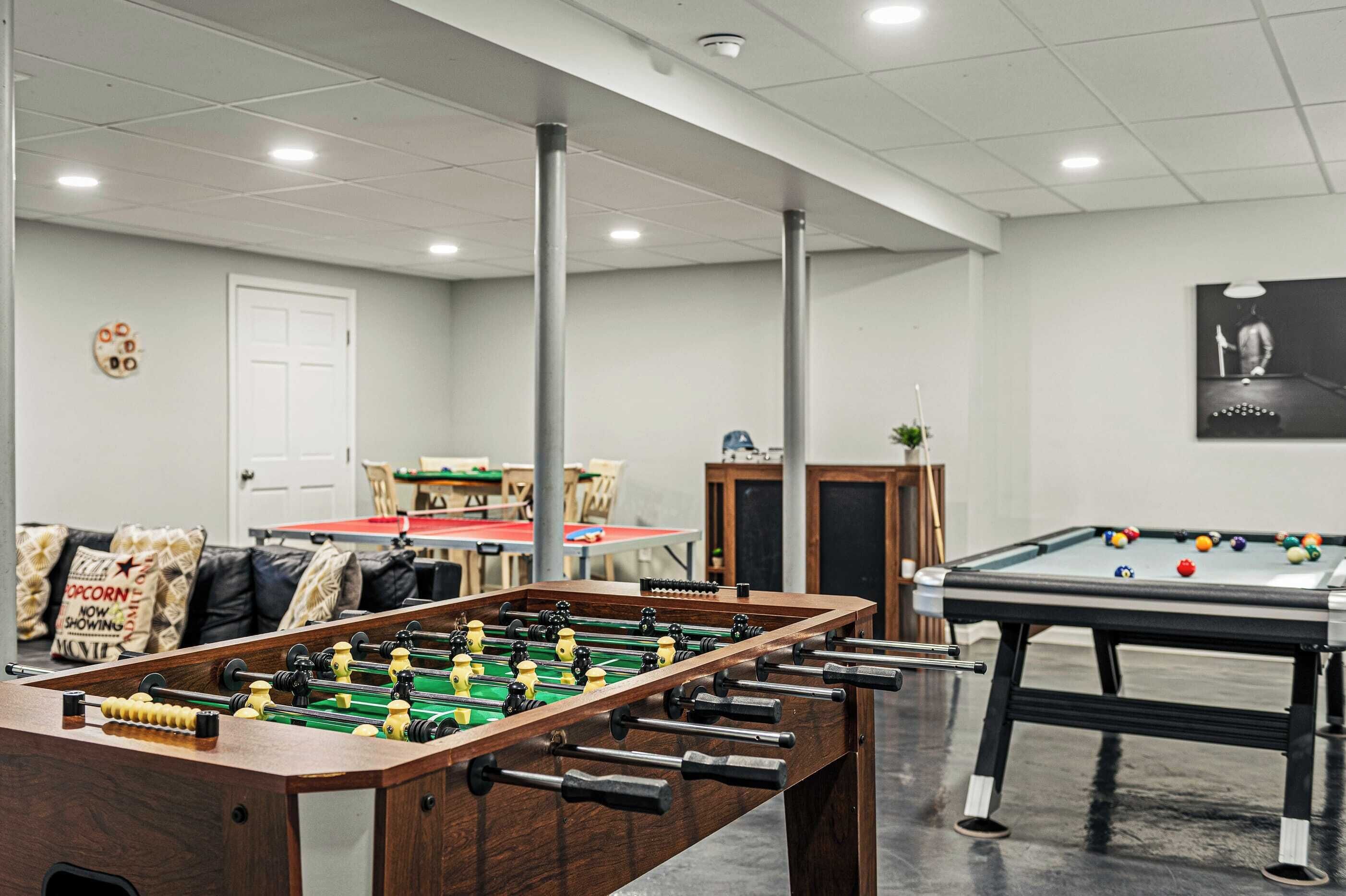A room with a foosball table, a pool table, and a billiards table. There are chairs and a table in the background.