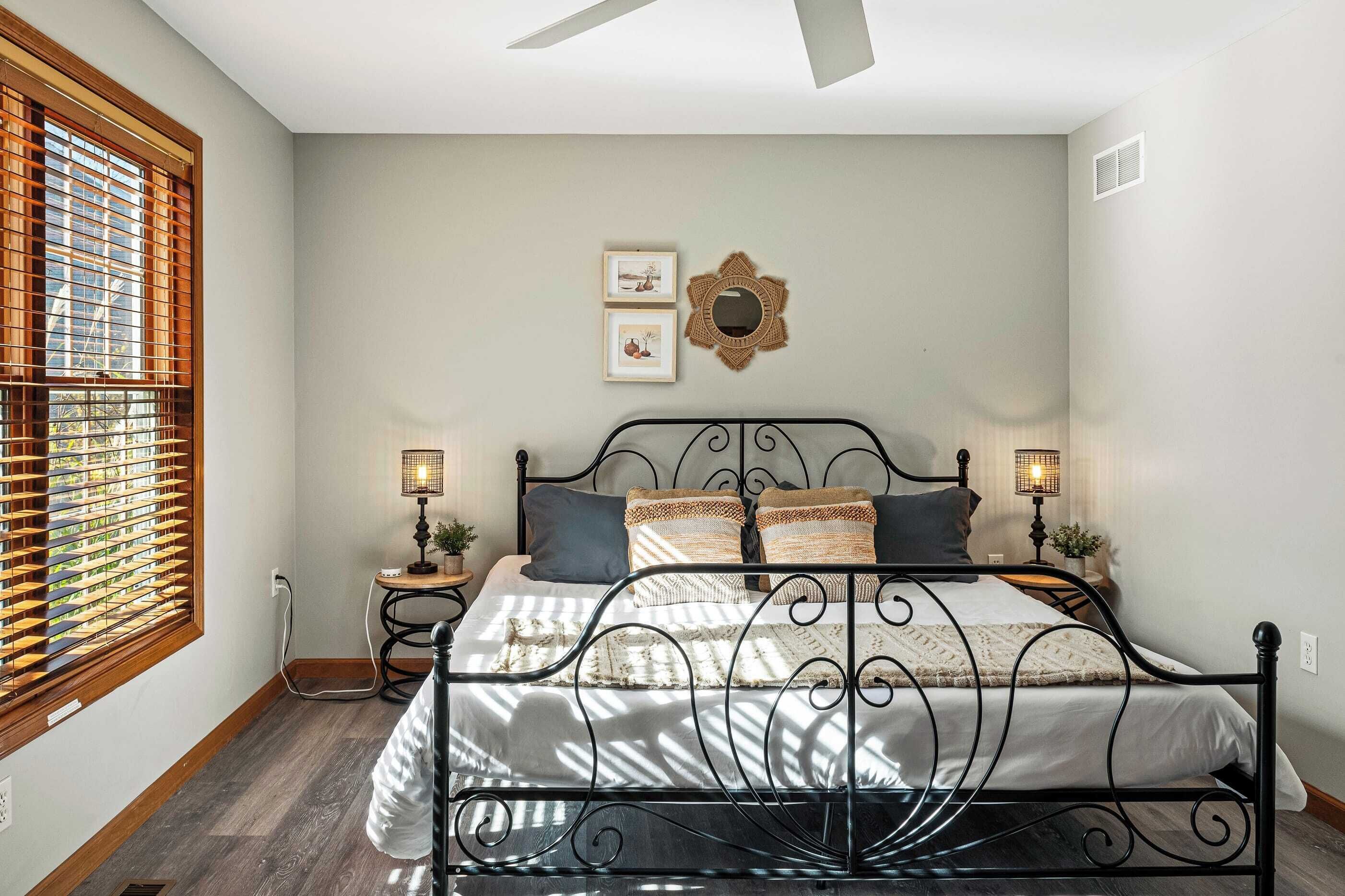 Une chambre à coucher comprenant un lit, deux tables de chevet, un miroir fixé au mur et une fenêtre avec des stores.