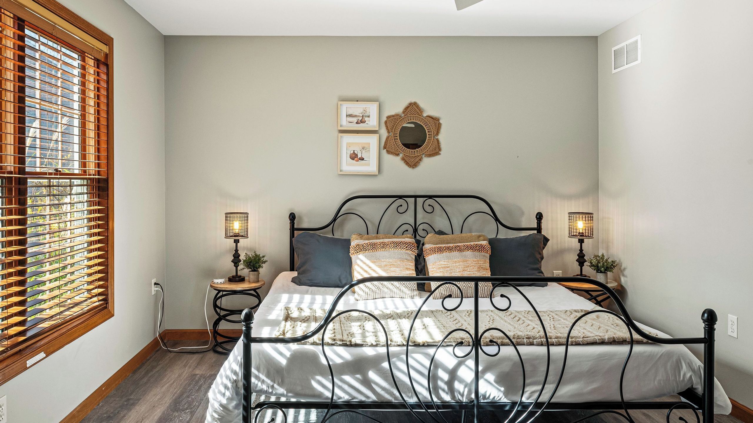 A bedroom with a bed, two bedside tables, a wall-mounted mirror, and a window with blinds.