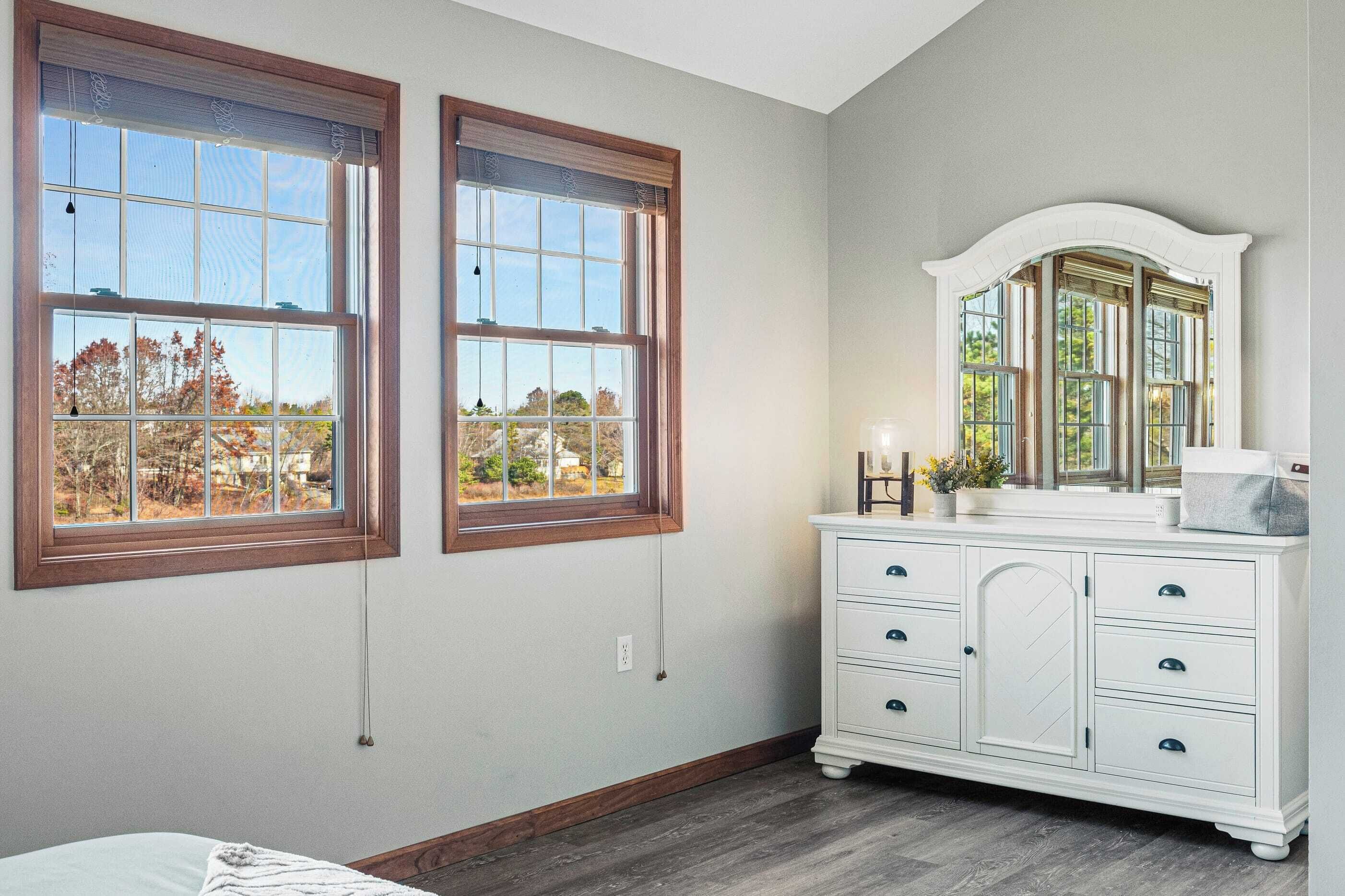 A bedroom with two large windows, a white dresser, and a mirror.