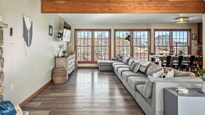 A spacious living room with a large gray sectional sofa, wooden beams on the ceiling, and a view of the outdoors through large windows. - Laker's Pointe by Avantstay Lakefront Private Peninsula Hot Tub, Game Room (Long Pond)